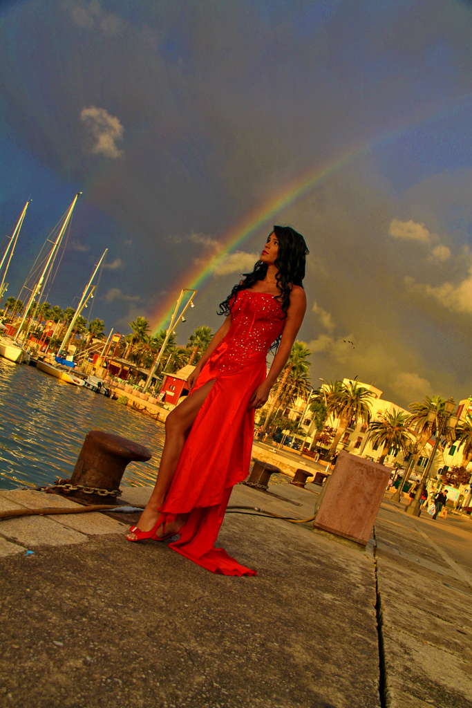 Model in a red dress on a marina pier beneath a rainbow